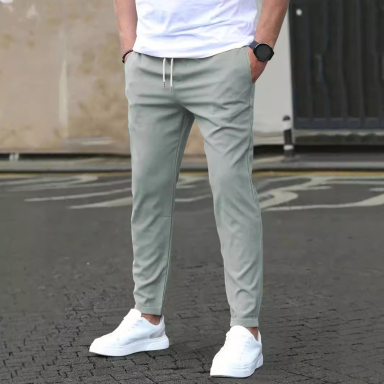 Hombre de pie con camiseta blanca y pantalones grises, calza zapatillas blancas.